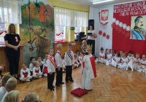Na tle biało czerwonej dekoracji stoi dziewczynka przebrana za Polskę oraz trzech chłopców przebranych za zaborców.